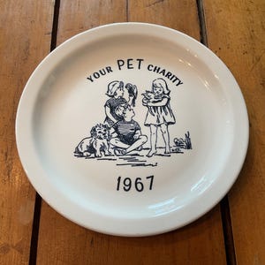 May include: White ceramic plate with a black illustration of children with pets and the text "YOUR PET CHARITY 1967". The plate has a wide rim and is set against a wooden surface.