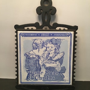 May include: Antique black cast iron trivet with a blue and white tile insert. The tile features an illustration of two children sharing candy, with the text "Home Made Candies" and "Watkins' 1912 Almanac". The trivet has a decorative handle.