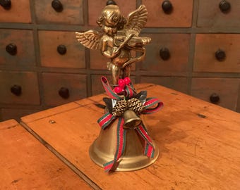 May include: A brass bell featuring a cherub playing a violin on top. The bell is decorated with a red and green striped ribbon, two small pinecones, and two red berries. The metal has a polished finish and a flared base design.