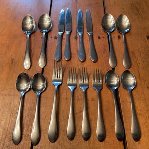 May include: A collection of stainless steel silverware, including spoons, forks, and knives, arranged on a wooden surface. The silverware has a simple, classic design, reflecting a traditional aesthetic. The arrangement suggests a formal dining setting.