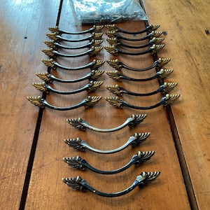 May include: A collection of decorative cabinet pulls. The pulls have a black finish with gold-colored leaf accents. Some pulls are silver-colored. A small bag of screws is visible in the background. The pulls are arranged on a wooden surface.