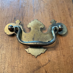 Peut inclure: Poignée de tiroir en laiton antique avec une poignée incurvée et une plaque arrière décorative. La quincaillerie présente une finition ternie, suggérant l'âge et l'utilisation. Idéale pour la restauration de meubles anciens.