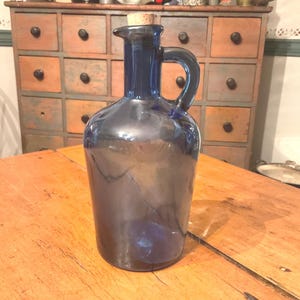 May include: A dark blue glass decanter with a handle and a cork stopper. The decanter is cylindrical with a rounded base and a spout. It sits on a wooden table. The background includes a wooden cabinet.