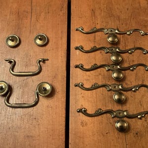 May include: A collection of antique brass cabinet hardware, including knobs and pulls. The knobs are round with a decorative edge, while the pulls feature a curved design. The hardware is arranged on a wooden surface, showcasing its vintage aesthetic.