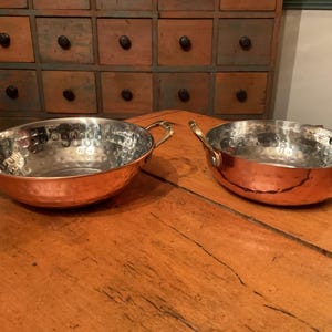 May include: Two copper-colored metal bowls with brass handles sit on a wooden table. The bowls have a hammered texture on the inside. The bowls are round and have a shiny, reflective surface.