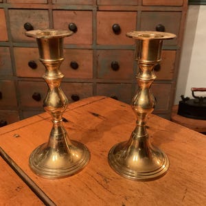 May include: Two antique brass candlesticks with a classic design. The candlesticks have a wide base, a slender stem, and a flared top. They are displayed on a wooden surface, with a vintage cabinet in the background.