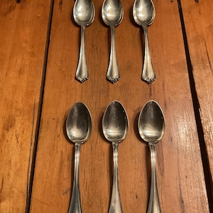May include: Six silver spoons arranged on a wooden surface. The spoons have a classic design with elongated handles and decorative detailing at the base of the bowl. The spoons are made of a shiny, reflective metal.