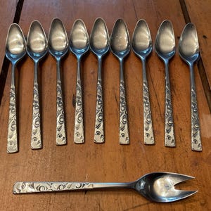 May include: A collection of nine silver spoons and a serving fork with patterned handles. The spoons are in a row, and the fork is beneath them. The handles feature an embossed design, suitable for serving.