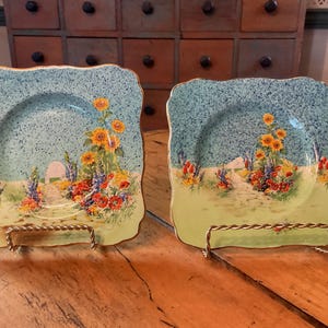 May include: Two square decorative plates with a floral design. The plates have a light blue speckled background with a garden scene featuring sunflowers and other colorful flowers. The plates are displayed on gold-colored stands.