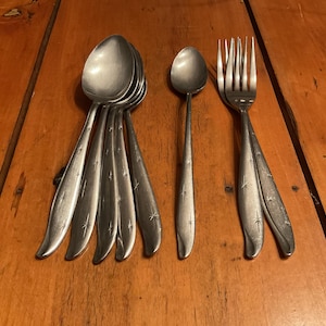 May include: A collection of vintage silverware, including spoons and a fork, arranged on a wooden surface. The spoons have a starburst design on the handles. The fork and spoons are made of a silver-colored metal.