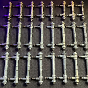 May include: A collection of twenty-one antique metal door handles. Each handle is a light gray color with a vertical bar design and small brass-colored accents. The handles are arranged in a grid pattern on a dark surface.