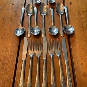 May include: A collection of vintage silverware, including spoons, forks, and knives, arranged on a wooden surface. The silverware has a silver finish with a decorative leaf pattern on the handles. The spoons are of various sizes, and the forks and knives are placed below.