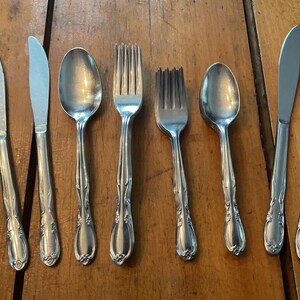 May include: A set of vintage silverware, comprising knives, spoons, and forks, displayed on a wooden surface. The silverware features decorative floral patterns on the handles. The knives have a curved blade, and the spoons have a rounded bowl.