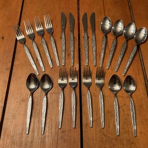 Peut inclure: Un ensemble de couverts en acier inoxydable, comprenant des fourchettes, des couteaux et des cuillères, disposés sur une surface en bois. Les couverts présentent un motif floral décoratif sur les manches.