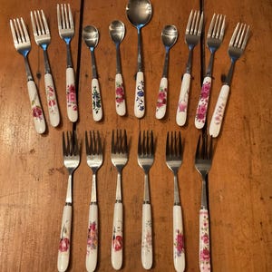 May include: A collection of fifteen pieces of stainless steel cutlery featuring white ceramic handles decorated with various floral patterns, including roses and other blossoms. The set consists of nine forks, three small spoons, two medium spoons, and one large spoon.