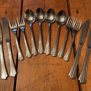 May include: A collection of stainless steel silverware, including spoons, forks, and knives, arranged on a wooden surface. The silverware has a simple, elegant design with a polished finish, reflecting the light. The set includes multiple pieces of each utensil.