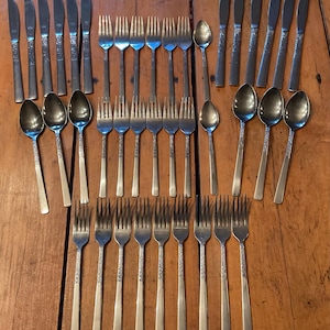 May include: A collection of vintage silverware, including knives, forks, and spoons, arranged on a wooden surface. The silverware has a decorative floral pattern on the handles. The set includes multiple place settings.