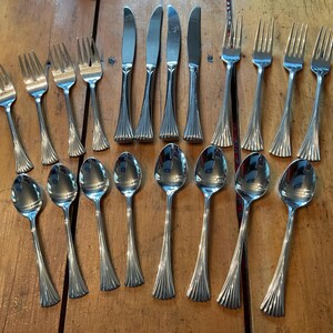 Peut inclure: Un ensemble de couverts en acier inoxydable, comprenant des fourchettes, des couteaux et des cuillères, disposés sur une surface en bois. Les couverts présentent un motif cannelé décoratif sur les manches. L'ensemble comprend 16 pièces.