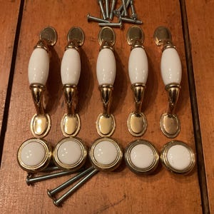 May include: A set of five arched cabinet handles and five matching round cabinet knobs, each featuring a white ceramic centre with polished gold-tone metal accents. The collection includes a group of metal mounting screws for installation.