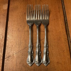 May include: Three silver forks with ornate handles are arranged vertically on a wooden surface. The forks have tines and decorative detailing on the handles. The wooden surface has a warm, brown tone.