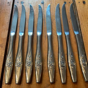 May include: A collection of eight silver butter knives with serrated blades. Each knife has a floral rose pattern on the handle. The knives are displayed on a wooden surface.