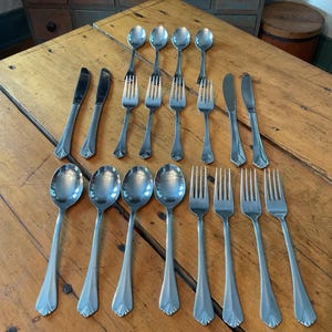 May include: A set of stainless steel cutlery, comprising spoons, forks, and knives, displayed on a wooden table. The cutlery features a straightforward, elegant design with a decorative edge. The table is crafted from light-coloured wood.