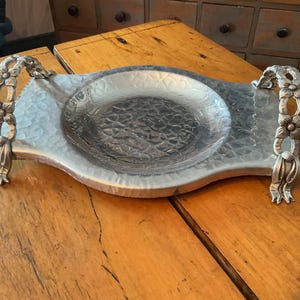 May include: Silver-toned metal serving tray with a hammered texture and floral handles. The tray has a central, slightly recessed area. The handles are adorned with flower-shaped details. A decorative piece for serving or display.