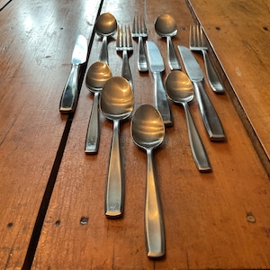 May include: A collection of stainless steel silverware, including spoons, forks, and knives, arranged on a wooden table. The utensils have a simple, modern design and reflect light. The table has a warm, natural wood tone.