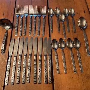May include: A collection of vintage cutlery, including forks, knives, spoons, and a ladle, arranged on a wooden surface. The cutlery has a decorative pattern on the handles and a silver finish. The set includes various sizes of spoons.