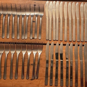 May include: A collection of stainless steel forks and knives arranged on a wooden surface. The forks are aligned in rows, with tines facing up. The knives are positioned parallel to each other, with blades facing the same direction. A complete set for dining.