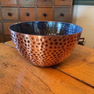May include: A copper bowl with a hammered texture. The bowl has a wide, rounded shape and a dark interior. The exterior is a warm copper color with a pattern of small, evenly spaced indentations. The bowl sits on a wooden surface.