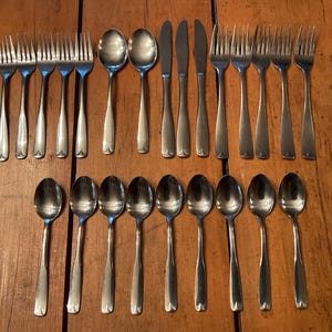 May include: A collection of stainless steel silverware, including forks, spoons, and knives, arranged on a wooden surface. The silverware has a simple, elegant design, with a smooth, polished finish. The forks, spoons, and knives are all the same style.