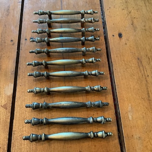 May include: A collection of ten antique brass drawer pulls, arranged on a wooden surface. Each pull has a central, elongated handle with decorative, rounded ends. The brass has a slightly aged patina.