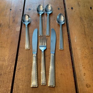 May include: A set of stainless steel silverware with gold-colored trim, including spoons, knives, and a fork, arranged on a wooden surface. The spoons are at the top, knives and fork in the middle, and spoons at the bottom.