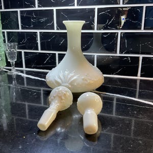 May include: A collection of off-white glass items, including a vase with a bulbous base and two bottle stoppers. The vase has a long neck and a decorative floral pattern. The stoppers have a round top and a cylindrical base. The items are on a black surface.
