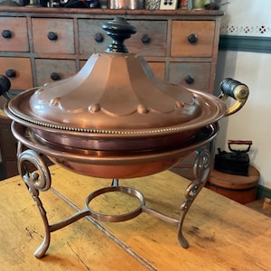 May include: A copper chafing dish with a domed lid and ornate handles. The dish sits on a decorative stand with scrolled legs. The lid has a black finial and a beaded edge. The chafing dish is a warm copper color.