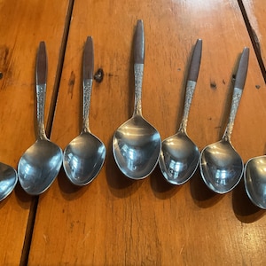 May include: A collection of seven spoons with silver bowls and wooden handles. The handles have a dark brown finish and are attached to the silver spoon bowls. The spoons are arranged on a wooden surface.