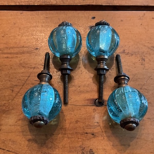 May include: Four decorative glass cabinet knobs. Each knob features a ribbed, light blue glass orb with a bronze-colored base and screw. The knobs are arranged on a wooden surface.
