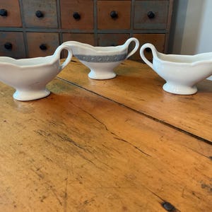 May include: Three white ceramic gravy boats with handles and spouts are arranged on a wooden table. One boat has a blue and white patterned band around the middle. The boats are oval-shaped with a pedestal base.