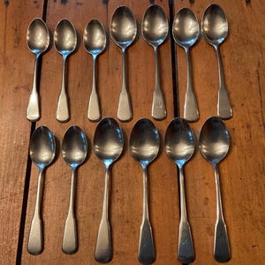May include: A collection of twelve silver spoons arranged on a wooden surface. The spoons have a classic design with oval bowls and rectangular handles. The spoons are displayed in two rows of six.