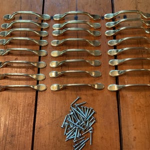 May include: A collection of twenty-one polished brass cabinet pulls arranged in three vertical columns, each featuring a smooth, arched design with rounded ends. A pile of silver-toned metal mounting screws is positioned below the handles.