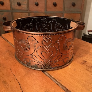 May include: A copper-colored metal container with a cylindrical shape and two handles. The container features a decorative pattern of birds and hearts created by punched-out holes. The interior is black, and the container sits on a wooden surface.