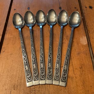 May include: Six silver spoons with long handles and ornate floral designs. The spoons are arranged in a row, with the bowls facing up. The spoons are on a wooden surface.