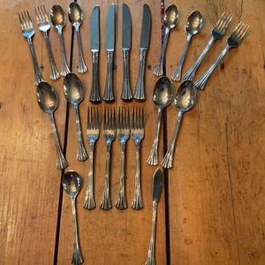 May include: A selection of vintage silverware, including forks, spoons, knives, and a butter knife, arranged on a wooden surface. The silverware has a silver finish with a decorative handle design. The arrangement suggests a formal dining setting.