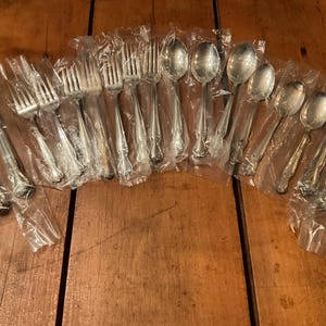May include: A collection of silver-colored flatware, including forks, spoons, and knives, individually wrapped in clear plastic. The silverware has ornate handle detailing. The items are arranged on a wooden surface.
