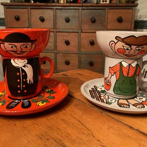 May include: Two ceramic sets featuring whimsical character designs. One set includes a red bowl, a black mug with a white scarf, and a matching plate. The other set has a white bowl, a mug with an orange shirt and green overalls, and a plate.