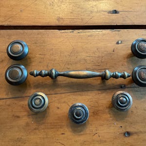 May include: A collection of antique brass hardware, including seven round knobs and a long, ornate handle. The hardware is arranged on a wooden surface, showcasing its aged patina and intricate detailing. Ideal for vintage furniture restoration.