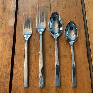May include: Four pieces of stainless steel silverware are arranged on a wooden surface. The set includes two forks and two spoons. The silverware has a hammered texture on the handles, and the spoons have a reflective surface.