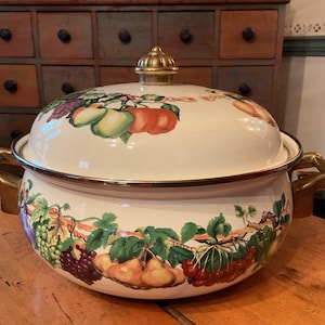 May include: A white enamel pot with a lid, featuring a colorful fruit design of apples, grapes, and pears. The pot has gold-colored handles and trim, and a gold knob on the lid. The pot is sitting on a wooden surface.
