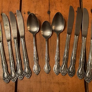 May include: A collection of vintage silverware, including knives and spoons, arranged on a wooden surface. The silverware has ornate, floral-patterned handles and shiny, reflective blades and bowls. The set includes various sizes of spoons and knives.
